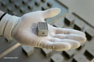A small square silver metal part being held in the palm of a hand with a white glove on
